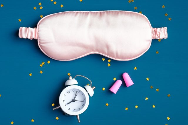 Sleep eye mask with alarm clock and ear plugs on dark blue background. Good night, sleep hygiene, insomnia treatment concept