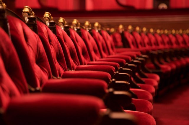 Traditional classically regal ornate rounded wood armed formal plush deep red velvet opera movie theater chairs in curved row with decorative gold molding in fancy carpeted venue